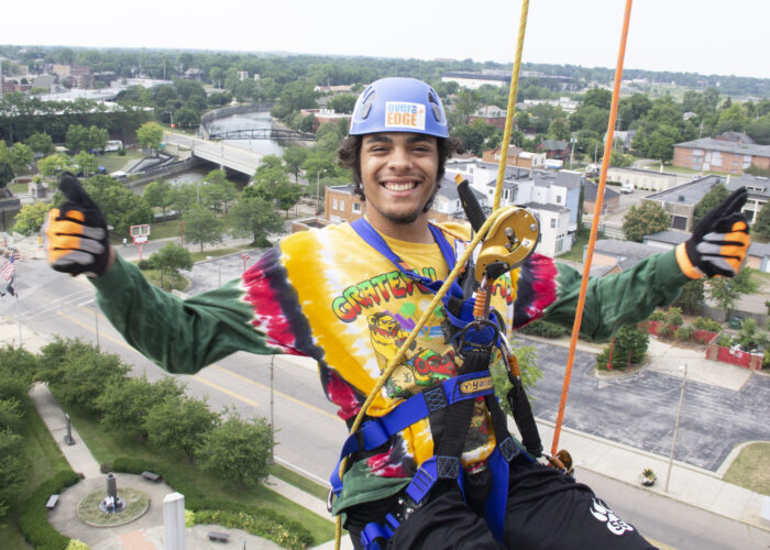 A smiling young man wear a tie die shirt and a helmet as he is suspended in a harness above the city