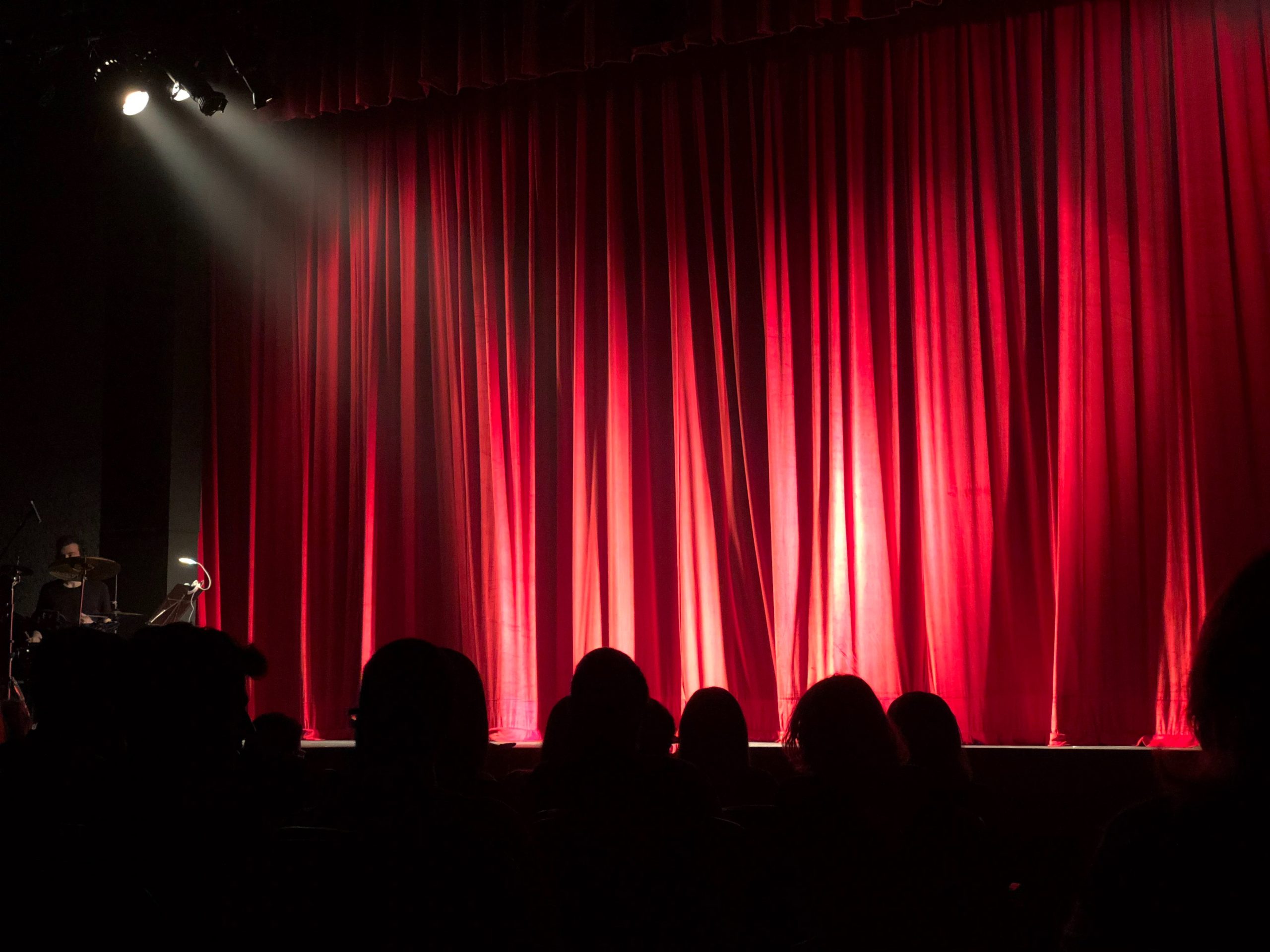 A stage with curtains closed is in front of the audience. in the background a drummer waits for their cue to play.