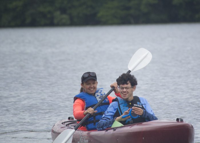 Two people sit in a kayak. The smiling woman in the back is paddling and in the front a boy wearing glasses smiles.