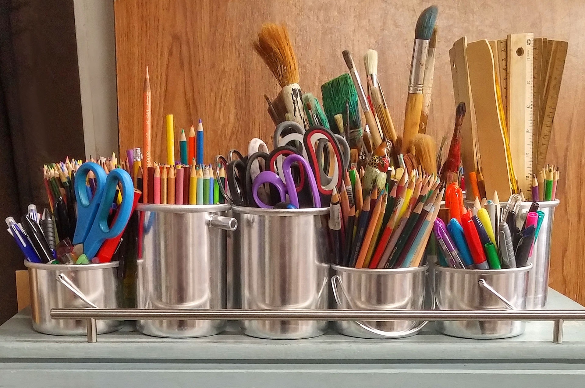 Colored pencils, markers, scissors, rulers, ink pens, and paintbrushes are shown in metal buckets.